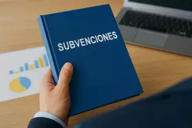 Mano sostiene un libro azul con "SUBVENCIONES" escrito. Detrás, gráficos y un portátil.