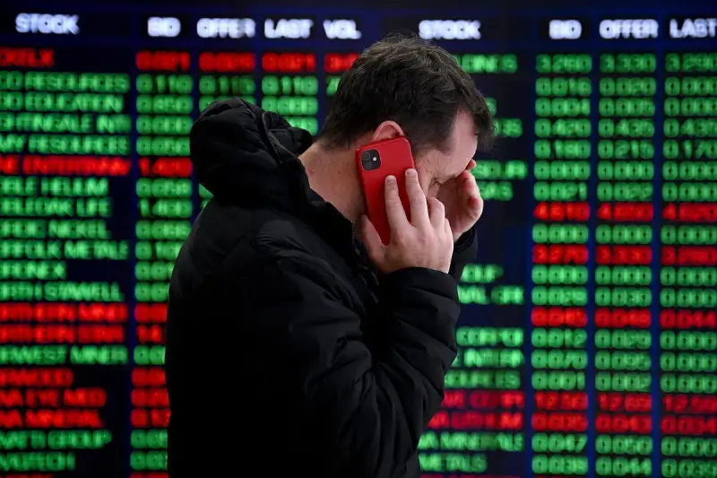 Hombre preocupado habla por teléfono frente a pantalla de bolsa de valores de Nueva York, con números rojos y verdes indicando un posible crack.